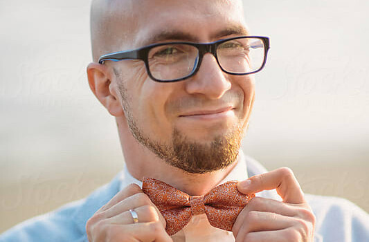 A man in a blue suit with glasses smiling into the camera holding onto his red bowtie.