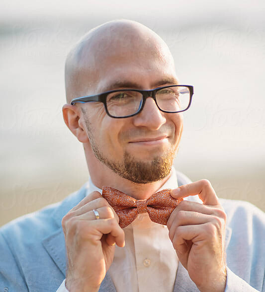 A man in a blue suit with glasses smiling into the camera holding onto his red bowtie.